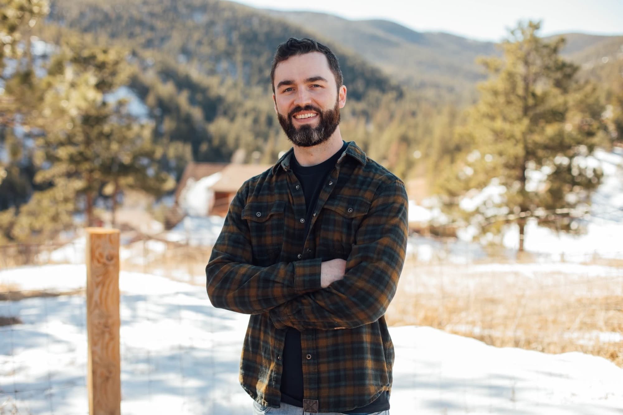 Ryan Williams standing outdoors in a flannel shirt, arms crossed, with snowy pines and mountains behind him